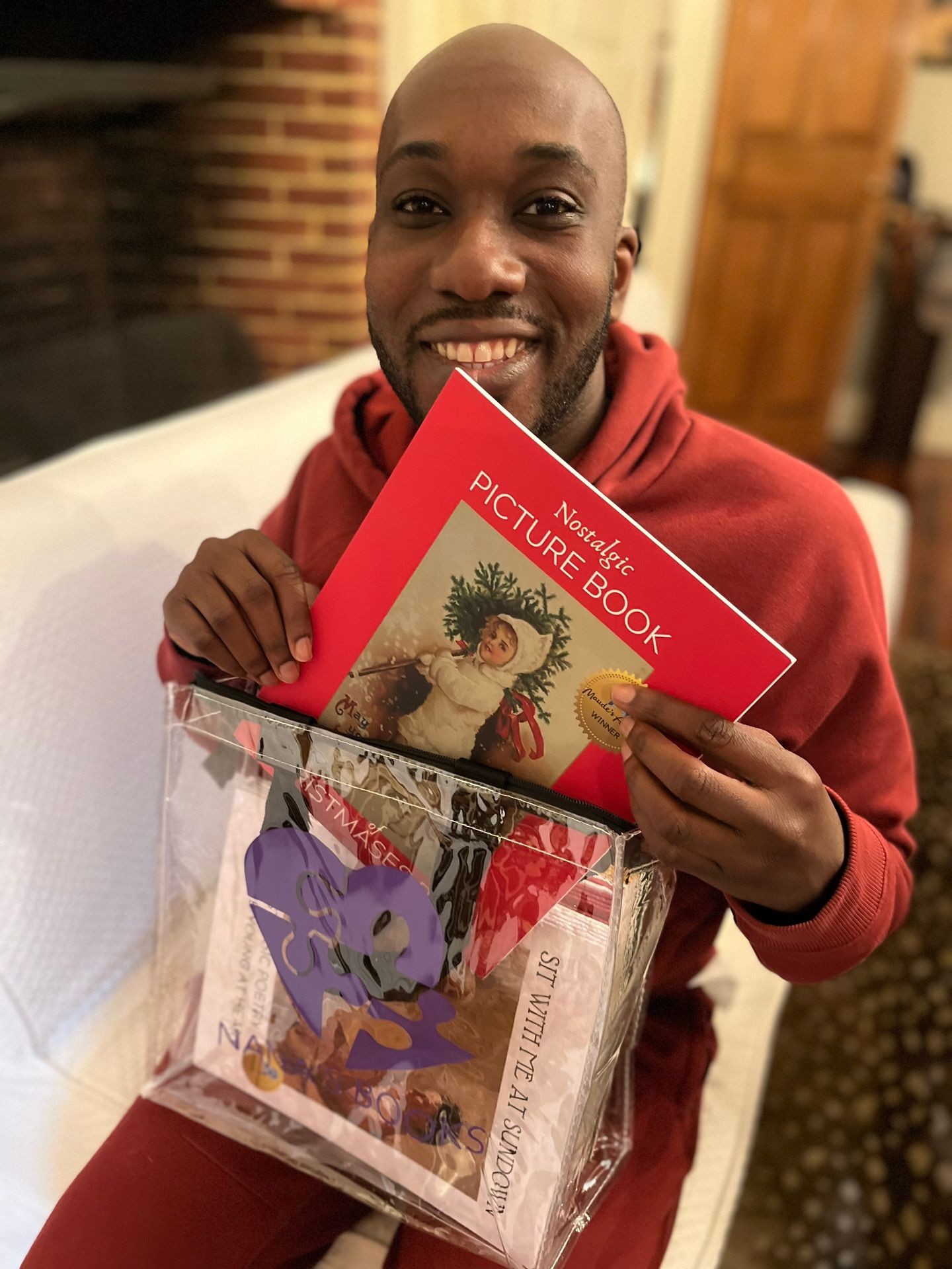 A young African American man in a red sweatshirt smiles as he unpacks a clear tote of Nostalgic Holiday books to share with a loved one in long term care. He is bald and sits contentedly near a brick fireplace.