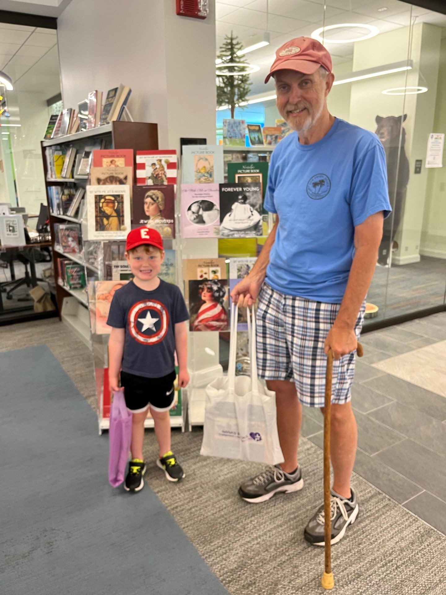 A man in plaid shorts and a baseball hat holds a cane and a tote bag filled with NANA’S BOOKS designed to meet the needs of people living with dementia. He stands alongside his grandson who holds a lavender tote bag of NANA’S BOOKS, as well, as they check out of the library together.