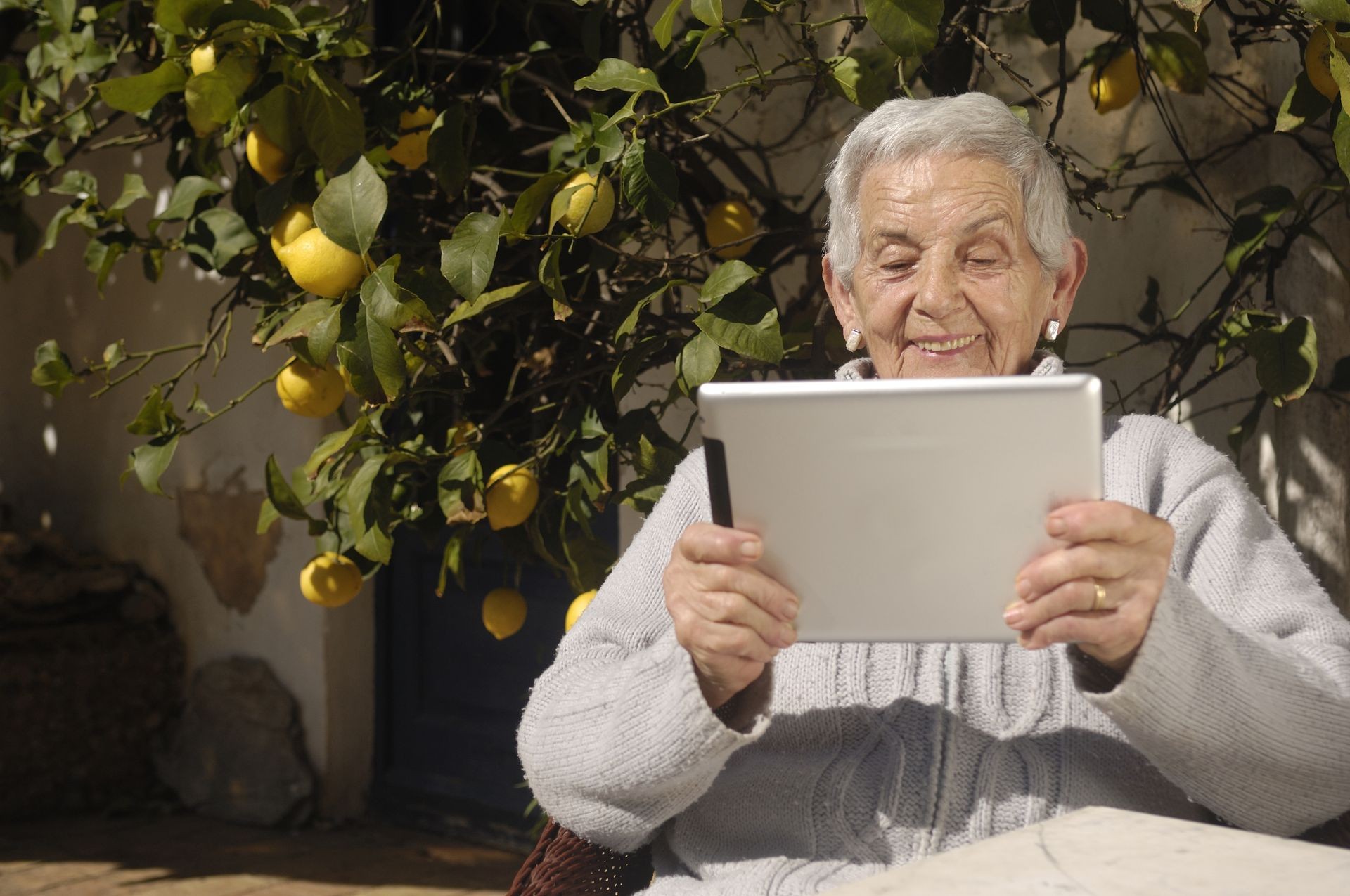  Smiling older woman sitting next to a lemon tree and reading on a tablet.